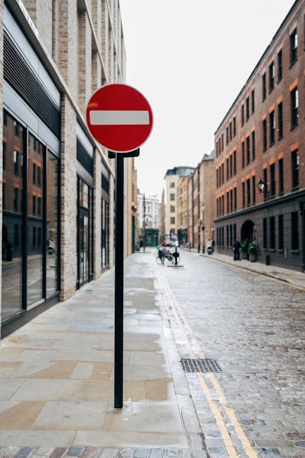 A narrow cobbled street in Shoreditch with tall, historic buildings on both sides, featuring stone façades and large windows. The street is mostly empty, with a few small barriers and a lamppost visi